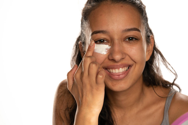 jonge donker gevilde vrouw het aanbrengen van een crème op haar lage oogleden op witte achtergrond - Foto, afbeelding