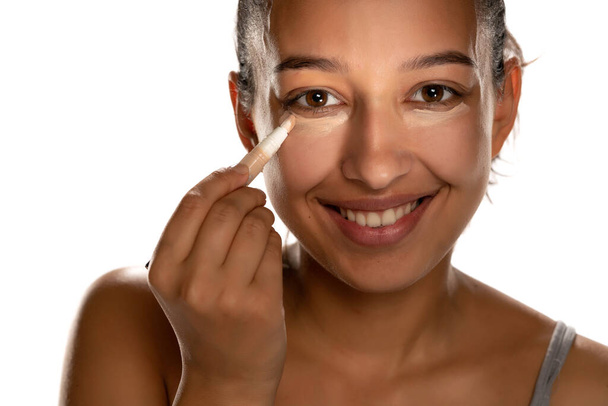 Young dark skinned woman applying concealer on her low eyelids on white background - Fotó, kép