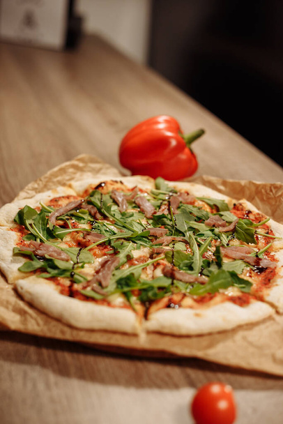 frische Pizza mit Kräutern, Thunfisch und Balsamico-Soße auf Esspapier auf einem Holztisch in einem gemütlichen Café, Paprika im Hintergrund - Foto, Bild