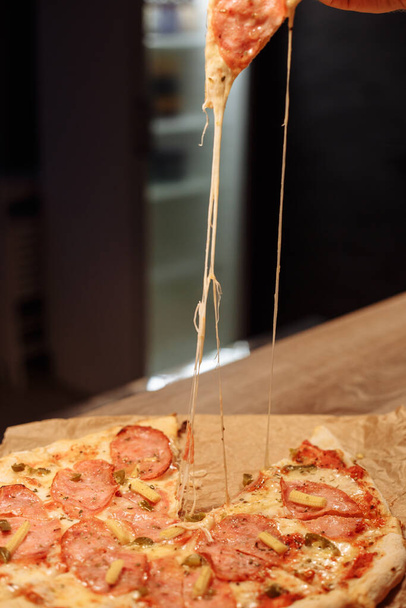 Το λιωμένο τυρί απλώνεται από μια φέτα πίτσα. Αρπάζοντας μια φέτα πίτσα με σαλάμι, μίνι καλαμπόκι και χαλαπένια. Πίτσα σε ξύλινο τραπέζι σε εστιατόριο - Φωτογραφία, εικόνα