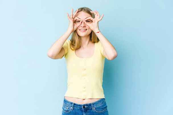 Young blonde woman isolated on blue background showing okay sign over eyes - Fotografie, Obrázek
