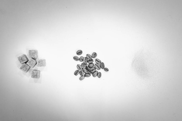 Schwarz-Weiß-Bild von Kaffeebohnen umgeben von Körnchen aus weißem Zucker und Würfeln aus braunem Zucker. - Foto, Bild