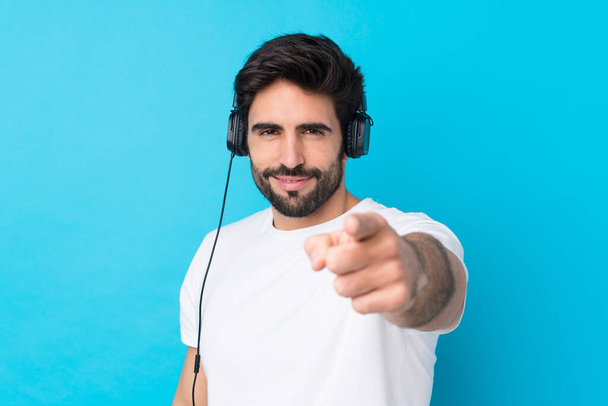 Nuori komea mies parta yli eristetty sininen tausta kuuntelee musiikkia ja osoittaa edessä - Valokuva, kuva