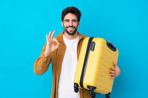 Młody przystojny mężczyzna z brodą na odizolowanym niebieskim tle na wakacjach z walizką podróżną i podejmowania OK znak - Zdjęcie, obraz