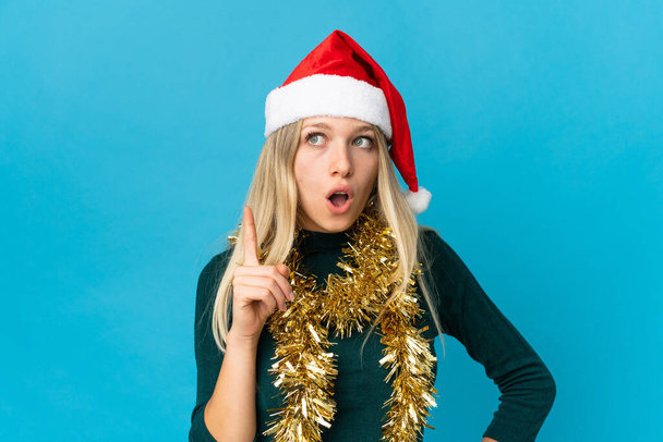 Жінка з різдвяним капелюхом ізольована на синьому фоні думаючи про ідею, спрямовану пальцем вгору
 - Фото, зображення