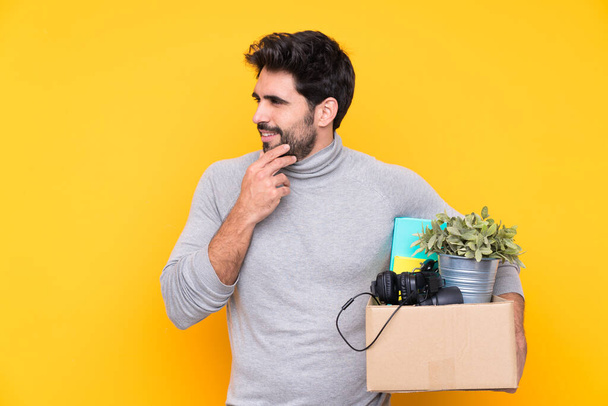 Young handsome man with beard making a move while picking up a box full of things over isolated background thinking an idea and looking side - Photo, Image
