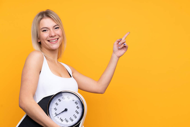 Young blonde Russian woman over isolated yellow background with weighing machine and pointing side - Photo, Image