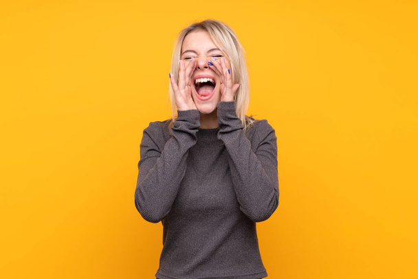 Young blonde woman over isolated yellow background shouting and announcing something - Foto, afbeelding