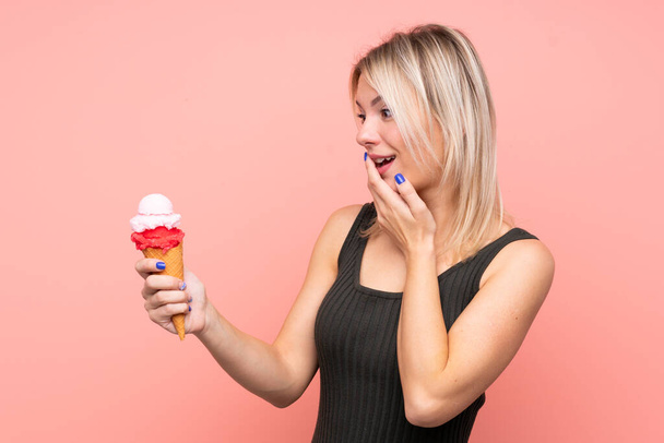 Молодая блондинка с мороженым из корнета на изолированном розовом фоне с удивлением и шокированным выражением лица - Фото, изображение