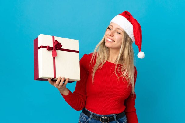 Woman with christmas hat holding a present isolated on blue background with happy expression - Photo, Image