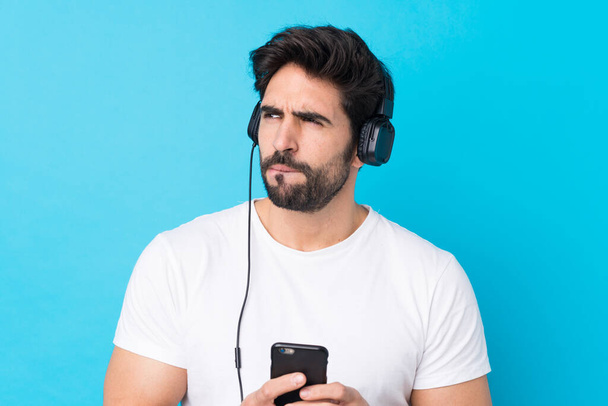 Молодий красивий чоловік з бородою над ізольованим синім фоном слухає музику з мобільним і мисленням
 - Фото, зображення
