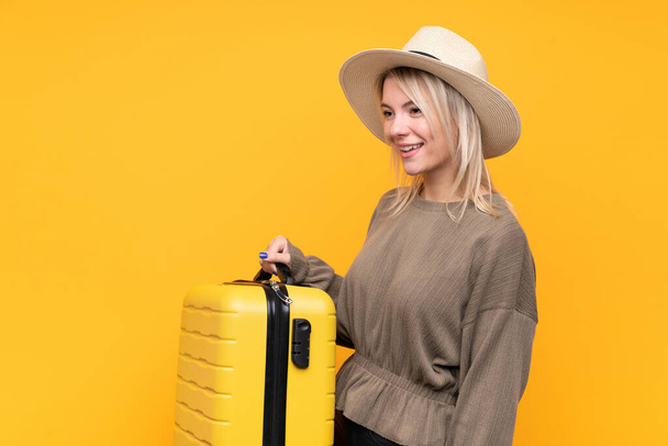Mujer rubia joven sobre fondo amarillo aislado en vacaciones con maleta de viaje y un sombrero - Foto, imagen