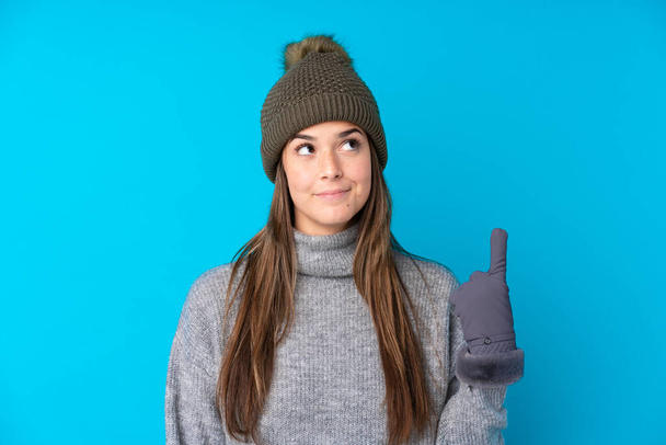 Chica adolescente con sombrero de invierno sobre fondo azul aislado apuntando con el dedo índice una gran idea - Foto, imagen