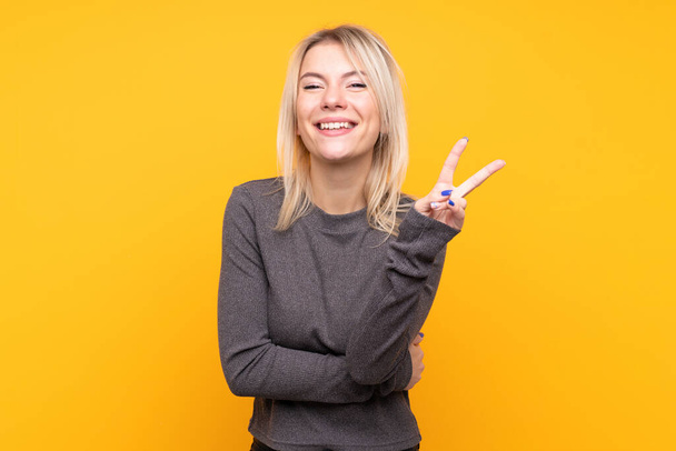 Young blonde woman over isolated yellow background smiling and showing victory sign - Photo, image