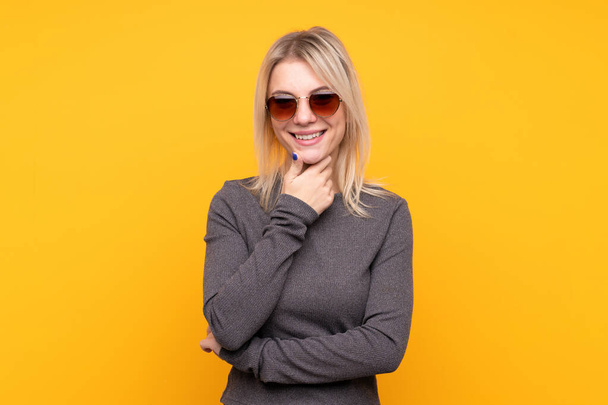 Jonge blonde vrouw over geïsoleerde gele achtergrond met bril en glimlachen - Foto, afbeelding