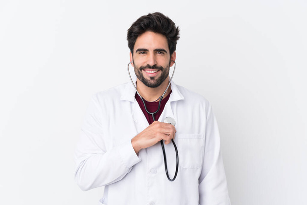 Молодой красивый мужчина с бородой на изолированном белом фоне в докторском платье и со стетоскопом - Фото, изображение
