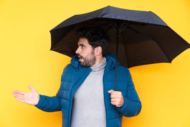 Homme tenant un parapluie sur fond jaune isolé avec expression faciale surprise - Photo, image