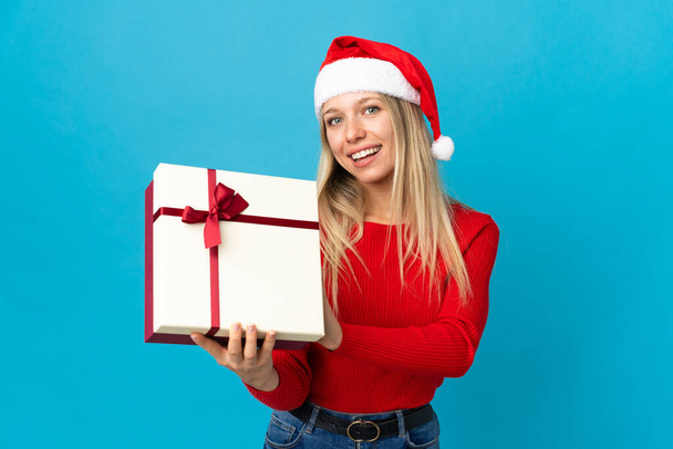Kobieta w świątecznym kapeluszu trzyma prezent na niebieskim tle ze szczęśliwym wyrazem twarzy - Zdjęcie, obraz