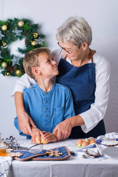Happy senior mature woman, grandmother and young boy, grandson cooking, kneading dough, baking pie, cake,  cookies. Family time in the cozy kitchen. Seasonal winter Christmas activity at home. - Photo, image
