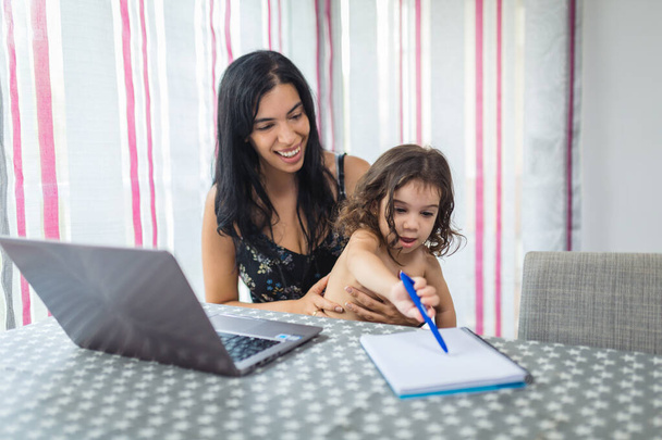 Latino anya teleworking egy laptop az ebédlőben, miközben ő a 2 éves lánya boldogan ül az ölében festmény egy notebook. - Fotó, kép