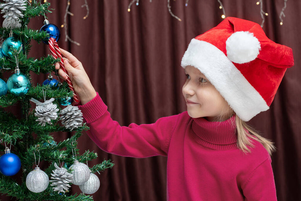 A cute Caucasian blonde girl in a red sweater and a Christmas cap hangs Christmas candy on a decorated Christmas tree. Christmas and New Year concept - Фото, изображение