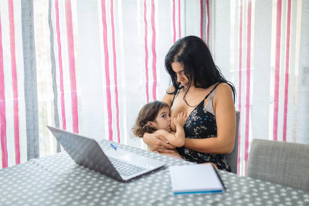 mãe teletrabalho na mesa da sala de jantar em um laptop enquanto amamenta sua menina que está sentada em seu colo - Foto, Imagem