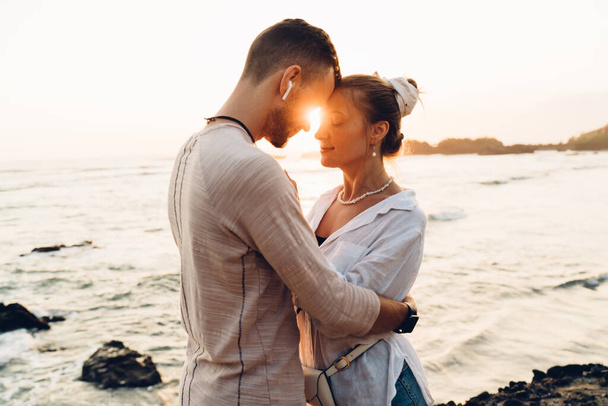 Side view of romantic male and female travelers holding each other while standing on stony coast near rippling sea during sunset - 写真・画像
