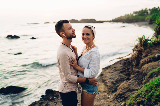 Cheerful man with wireless earphones standing on sandy shore next to girlfriend while having romantic holidays together during summer vacation in nature - Foto, Imagem