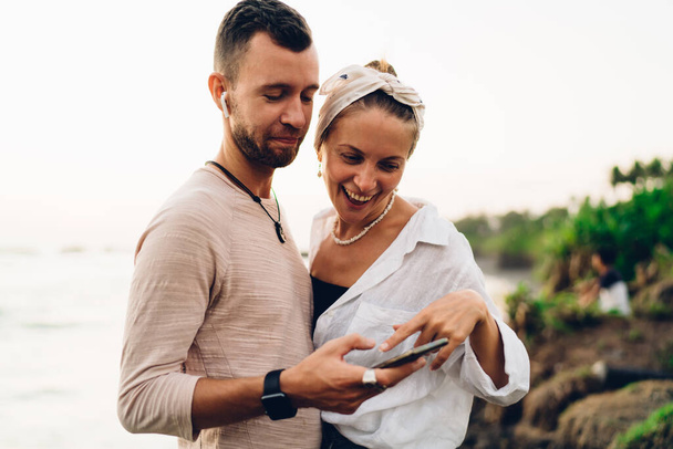 Smiling male and woman in casual clothes standing close to each other while watching funny video on cellphone on seashore - Фото, зображення