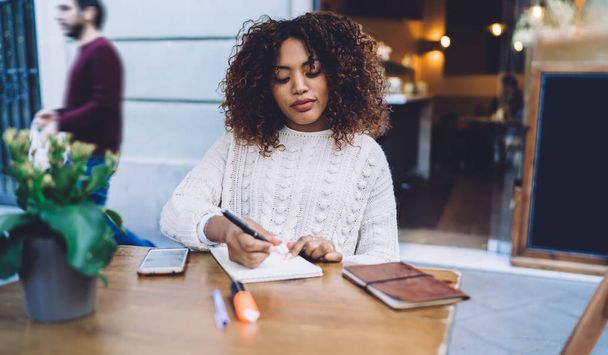 Positive African American female in casual clothes sitting at wooden table with smartphone and taking notes in copybook while resting in cafe - Фото, зображення
