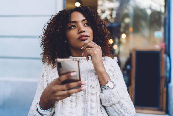 Pensive African American female in casual clothes sitting at wooden table and browsing mobile phone while resting in outdoor cafe - Photo, Image