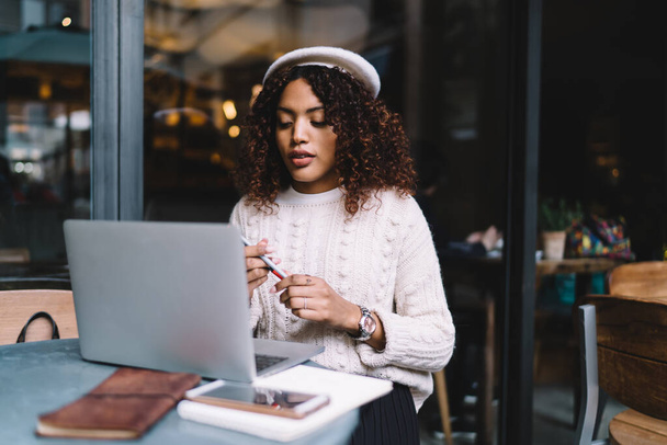 Young woman with curly hair in casual clothes sitting at table and using laptop for remote work in workspace of cafe - Foto, Imagem