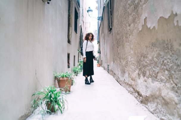 Young dark skinned female in trendy wear standing on street explore locations and destinations, beautiful 20s african american woman enjoying free time on urban settings in town on leisure - 写真・画像