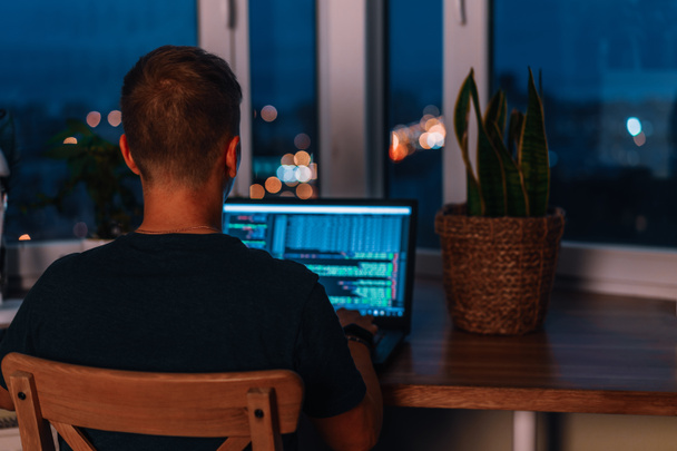 Mladý muž programátor kódování na notebooku ve tmě s výhledem na světla nočního města, barevné osvětlení v místnosti, domácí výzdoba - Fotografie, Obrázek