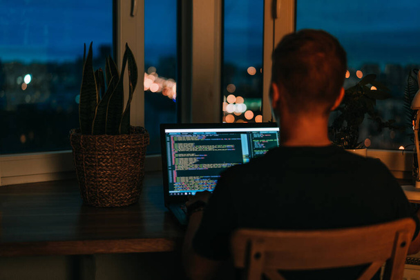 A young man programmer coding on a laptop in the dark with a view of the lights of the night city, color lighting in the room, home decor - Photo, Image