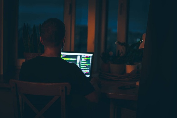 Mladý muž programátor kódování na notebooku ve tmě s výhledem na světla nočního města, barevné osvětlení v místnosti, domácí výzdoba - Fotografie, Obrázek