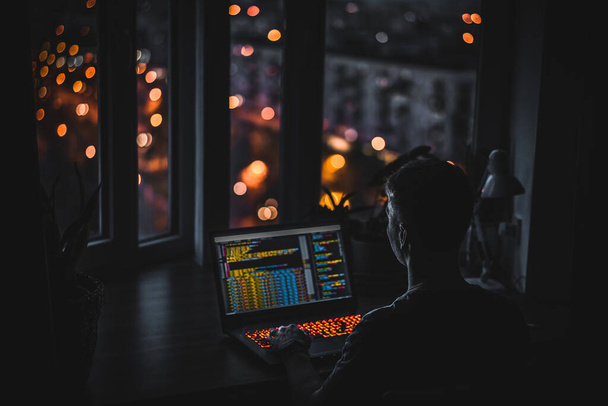 Un giovane programmatore codifica su un computer portatile al buio con una vista delle luci della città di notte, illuminazione a colori in camera, arredamento domestico - Foto, immagini