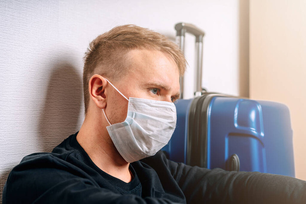 医療マスクの男はロシアのパスポートを保持し、空港で荷物の隣に座っています。コロナウイルスe中の旅行の概念 - 写真・画像