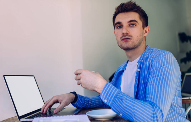 Portrait of skilled male graphic designer with tea cup sitting a desktop table with mockup laptop and posing, Caucasian freelancer using blank netbook with copy space area for internet advertising - Foto, Imagen