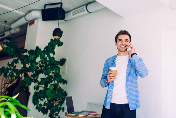 From below of joyful male in casual wear smiling and talking on mobile phone and holding cup of hot drink - Фото, изображение