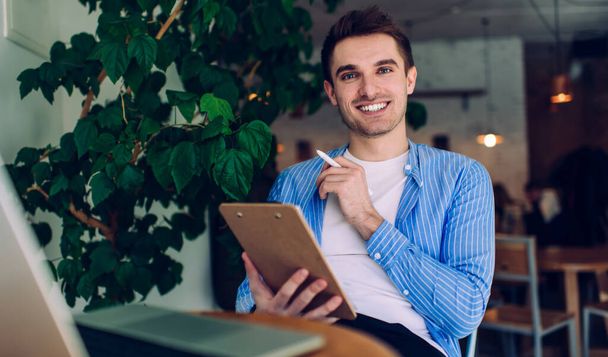 Smiling businessman in casual wear looking at camera and using laptop and taking notes while sitting at table and working on project in modern cafe - Φωτογραφία, εικόνα