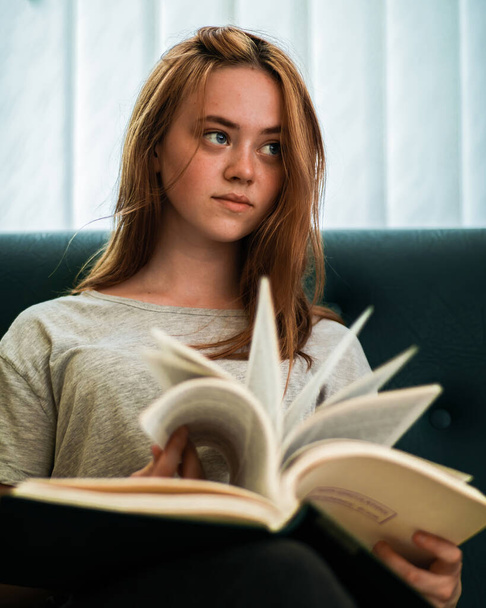 Μπλε μάτια κοκκινομάλλα κορίτσι με σοβαρή έκφραση του προσώπου κάθεται σε έναν καναπέ σε δημόσια βιβλιοθήκη και περιήγηση μέσα από ένα βιβλίο, ενώ κοιτάζοντας μακριά. - Φωτογραφία, εικόνα