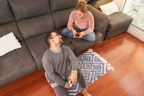 Vista dall'alto di una giovane coppia felice che si rilassa parlando tenendo in mano tazze di caffè e utilizzando il telefono sul divano insieme in soggiorno. - Foto, immagini