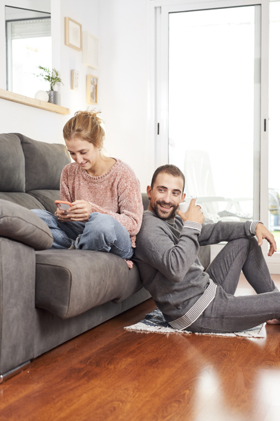 Feliz joven pareja relajándose hablando sosteniendo tazas de café y usando el teléfono en el sofá juntos en la sala de estar. - Foto, Imagen