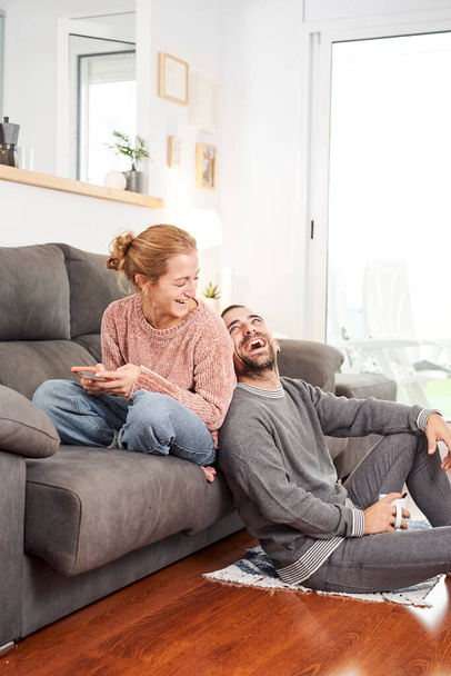 Šťastný mladý pár smích drží šálky kávy a pomocí telefonu na gauči spolu v obývacím pokoji. - Fotografie, Obrázek