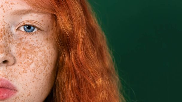 半顔の赤い髪の少女の肖像画で、肌にはそばかすがたくさんあり、カメラを覗き込む青い目、緑の背景 - 写真・画像