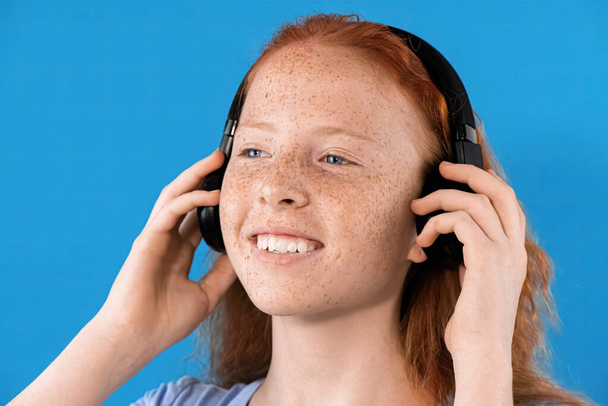 Retrato de una niña pelirroja sonriente con muchas pecas en la cara y ojos azules mirando hacia otro lado, escuchando música en auriculares, fondo azul - Foto, imagen