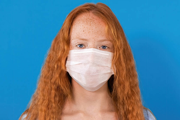 Portrait of a red-haired girl in a white medical mask with lots of freckles on the face and blue eyes looking into the camera, blue background - Foto, Bild