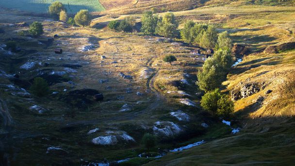 Natuur van Moldavië, vallei met stromende rivier en zeldzame bomen, rotsachtig land en zwarte vaten op de grond, gezaaid velden op de achtergrond - Foto, afbeelding