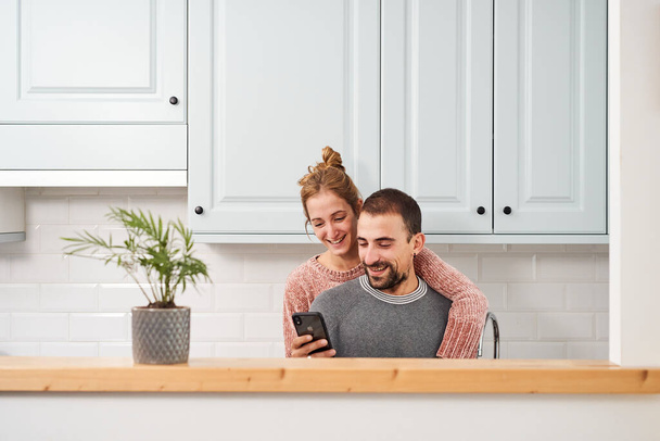Feliz pareja joven en la cocina en casa, usando el teléfono móvil y riendo. - Foto, imagen